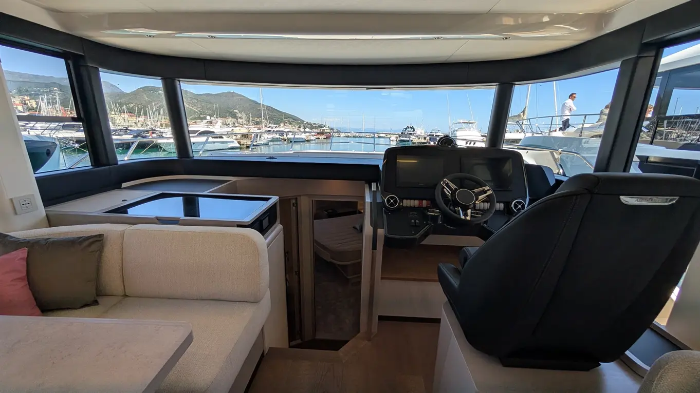 Interior view of a luxury boat's helm with steering wheel and controls, offering a panoramic marina view through large windows.